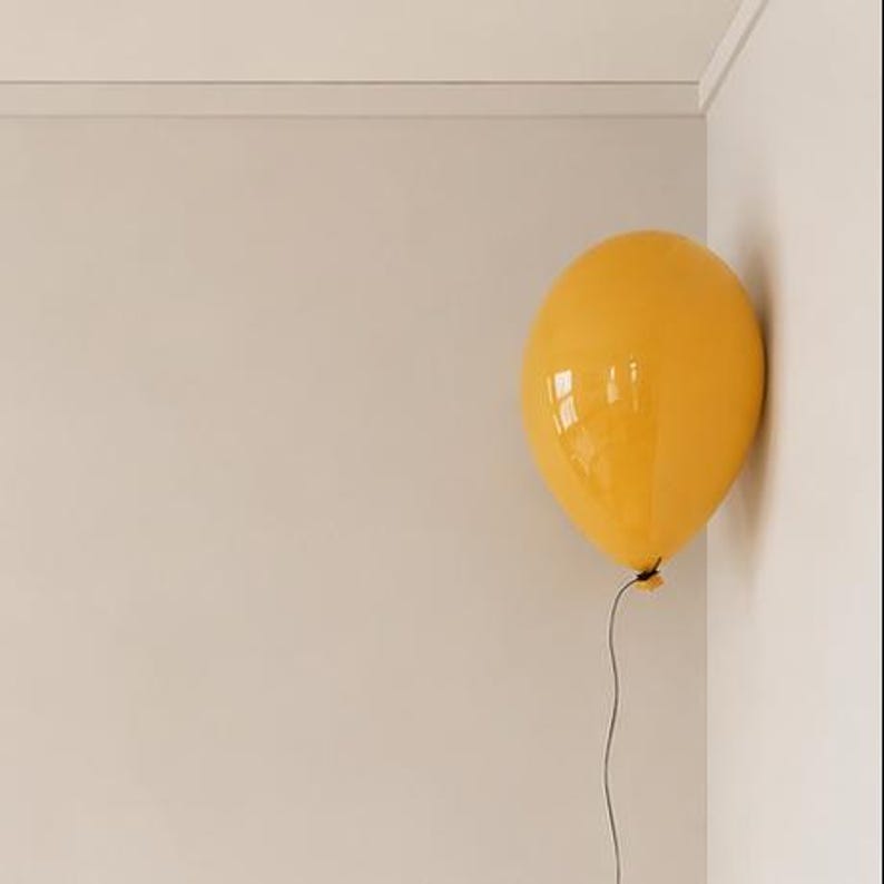 May include: A single, smooth, glossy yellow balloon floating in the corner of a room. The balloon is round and vibrant, featuring a thin, light-colored string hanging downwards from the tied knot at the base, against plain light-colored walls.