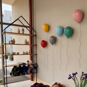 Puede incluir: Una estanter&iacute;a de metal negro con forma de casa y estantes de madera que exhibe varios objetos decorativos peque&ntilde;os y art&iacute;culos de felpa, junto a seis esculturas de pared brillantes con forma de globo en rosa, amarillo, rojo, gris, azul y verde.