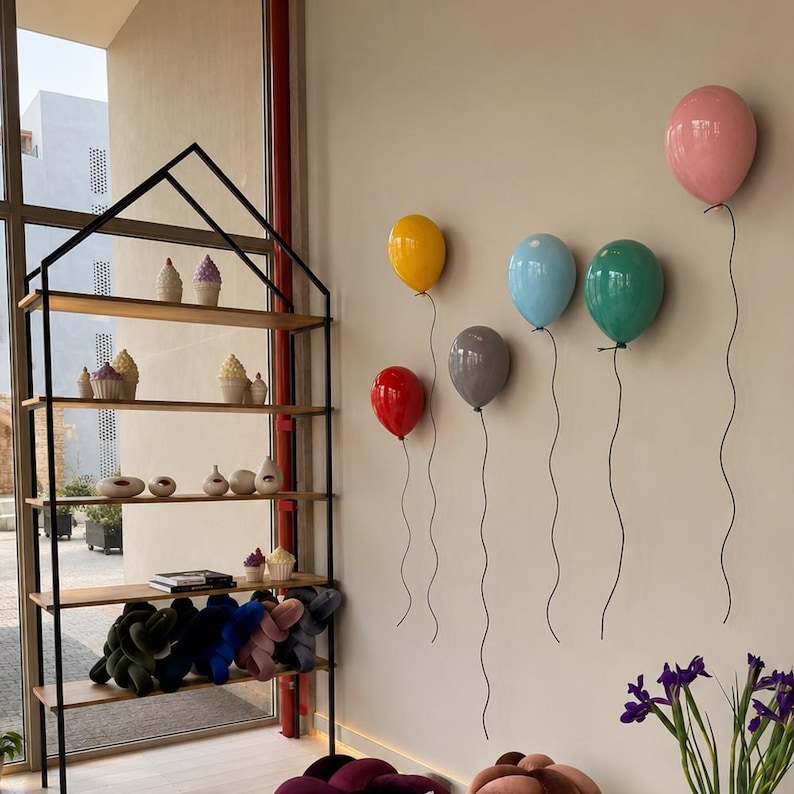 Puede incluir: Una estanter&iacute;a de metal negro con forma de casa y estantes de madera que exhibe varios objetos decorativos peque&ntilde;os y art&iacute;culos de felpa, junto a seis esculturas de pared brillantes con forma de globo en rosa, amarillo, rojo, gris, azul y verde.