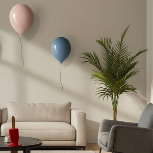 K&ouml;nnte beinhalten: Zwei ballonf&ouml;rmige Wanddekorationen, eine hellrosa und eine blaue, sind an einer neutralfarbenen Wand angebracht. Die Ballons haben eine gl&auml;nzende Oberfl&auml;che und sind an d&uuml;nnen, dunklen Schn&uuml;ren aufgeh&auml;ngt. Die Szene umfasst ein Sofa, einen Sessel und eine kleine Palme.