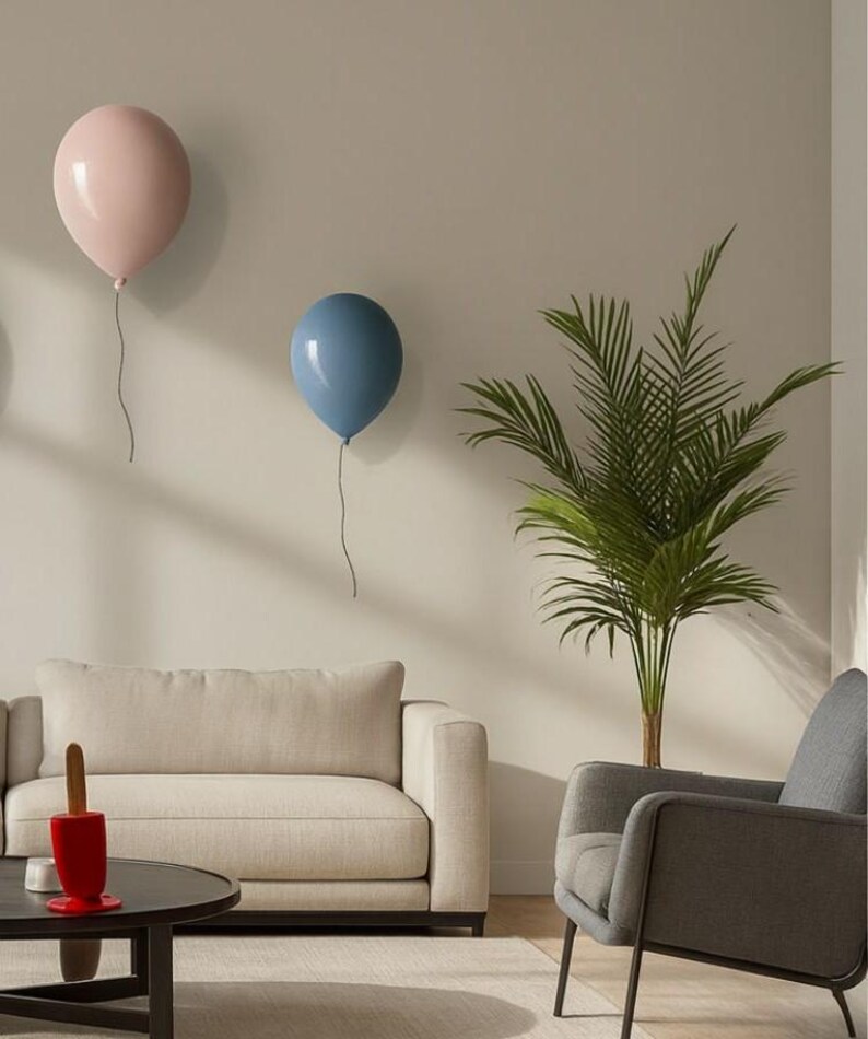 K&ouml;nnte beinhalten: Zwei ballonf&ouml;rmige Wanddekorationen, eine hellrosa und eine blaue, sind an einer neutralfarbenen Wand angebracht. Die Ballons haben eine gl&auml;nzende Oberfl&auml;che und sind an d&uuml;nnen, dunklen Schn&uuml;ren aufgeh&auml;ngt. Die Szene umfasst ein Sofa, einen Sessel und eine kleine Palme.