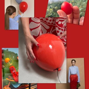 May include: A glossy red balloon-shaped object with a black string attached to the bottom. The surface is smooth and reflective, showing a bright highlight. The object is held by hands with red nail polish, set against a red background.