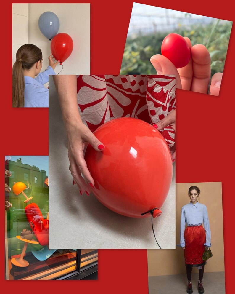 May include: A glossy red balloon-shaped object with a black string attached to the bottom. The surface is smooth and reflective, showing a bright highlight. The object is held by hands with red nail polish, set against a red background.