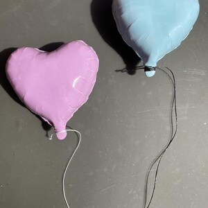Op de afbeelding: Twee hartvormige ballonnen, &eacute;&eacute;n lichtblauw en &eacute;&eacute;n lichtpaars, zijn aan elkaar gebonden met touwtjes. De ballonnen zweven tegen een grijze achtergrond.