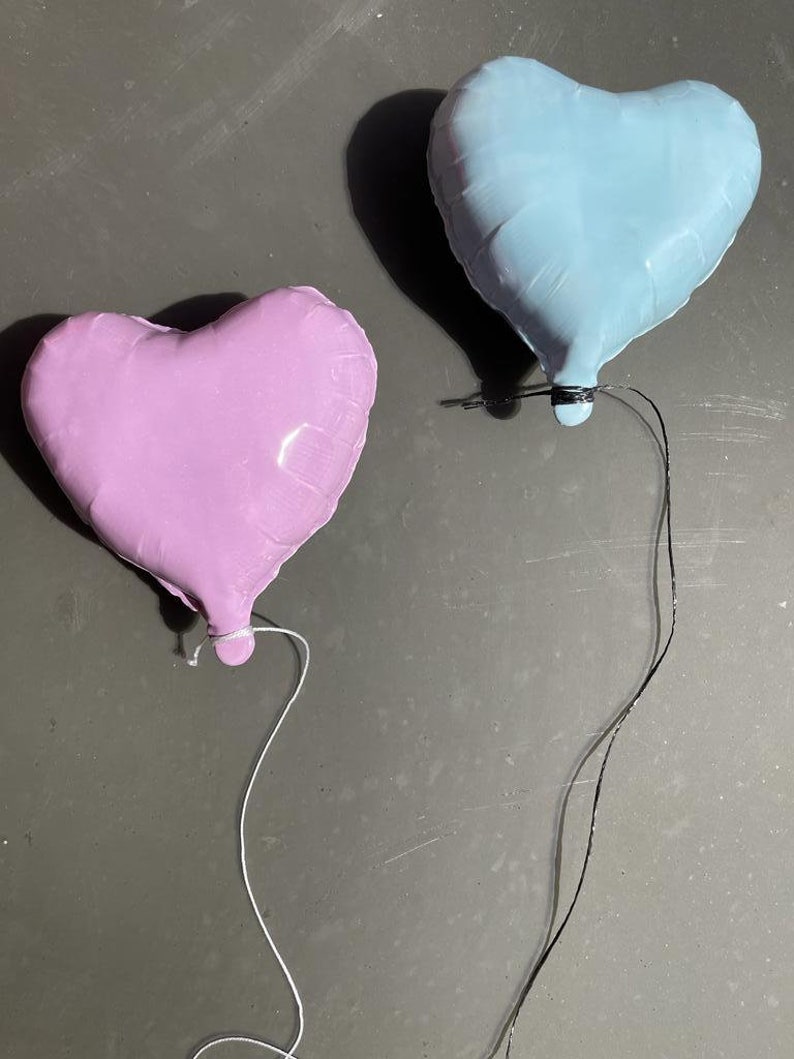 Op de afbeelding: Twee hartvormige ballonnen, &eacute;&eacute;n lichtblauw en &eacute;&eacute;n lichtpaars, zijn aan elkaar gebonden met touwtjes. De ballonnen zweven tegen een grijze achtergrond.