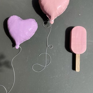 Op de afbeelding: Twee hartvormige ballonnen, &eacute;&eacute;n roze en &eacute;&eacute;n paars, zijn vastgebonden met wit touw. Een roze ijslolly op een houten stokje bevindt zich aan de linkerkant van de afbeelding.