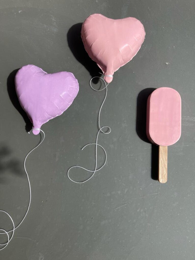Op de afbeelding: Twee hartvormige ballonnen, &eacute;&eacute;n roze en &eacute;&eacute;n paars, zijn vastgebonden met wit touw. Een roze ijslolly op een houten stokje bevindt zich aan de linkerkant van de afbeelding.