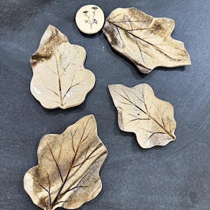 Puede incluir: Cuatro pequeños platos de cerámica en forma de hoja con un esmalte marrón y beige. Los platos están dispuestos sobre una superficie gris oscuro. Un pequeño azulejo de cerámica redondo con un diseño floral se encuentra en la esquina superior izquierda de la imagen.