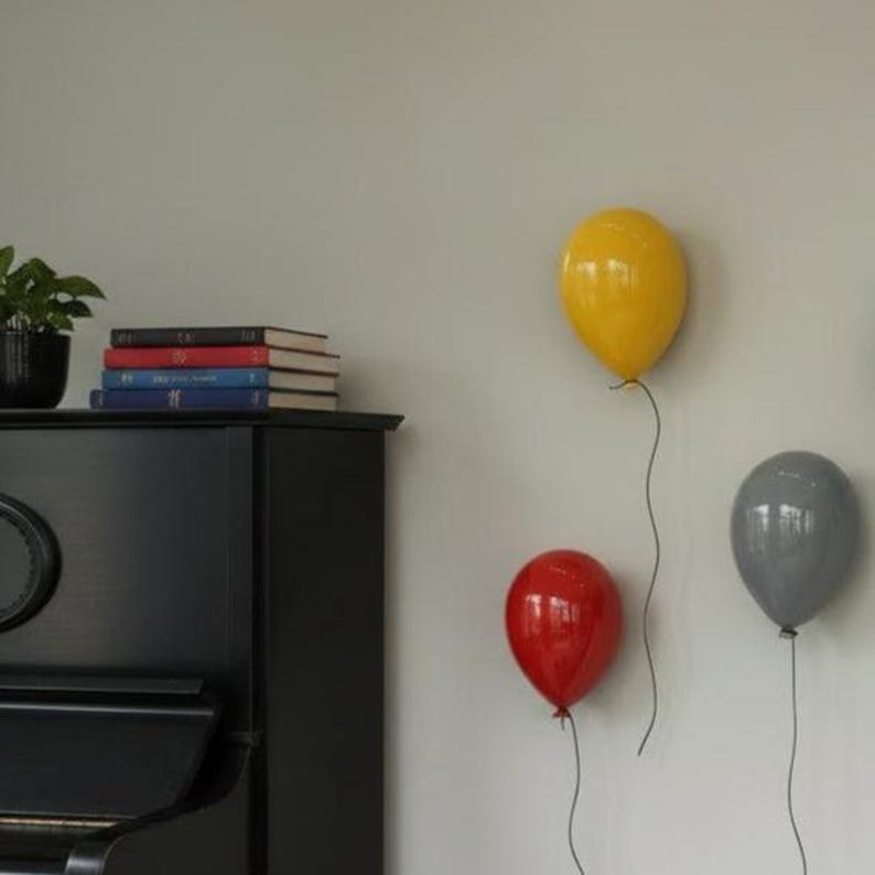 Puede incluir: Tres l&aacute;mparas de pared con forma de globo en colores amarillo, rojo y gris est&aacute;n montadas en una pared. Un piano negro se sit&uacute;a a la izquierda con una pila de cuatro libros encima. La l&aacute;mpara amarilla est&aacute; m&aacute;s alta, y las otras m&aacute;s abajo.