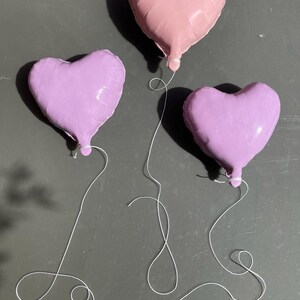 Op de afbeelding: Drie hartvormige ballonnen, &eacute;&eacute;n roze en twee paars, zijn aan elkaar gebonden met wit touw.
