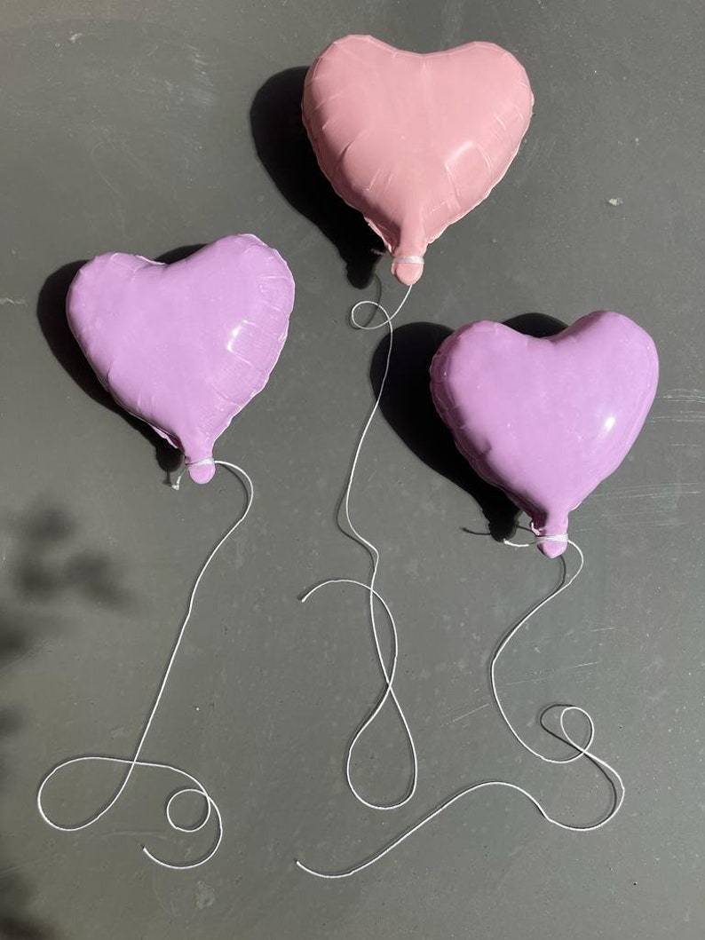 Op de afbeelding: Drie hartvormige ballonnen, &eacute;&eacute;n roze en twee paars, zijn aan elkaar gebonden met wit touw.