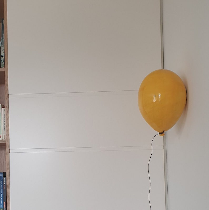 Puede incluir: Un aplique de pared con forma de globo amarillo y acabado brillante, montado directamente sobre una pared blanca. Un fino cable negro desciende desde la base del globo, que presenta un peque&ntilde;o detalle de nudo donde el cable se une a la l&aacute;mpara.