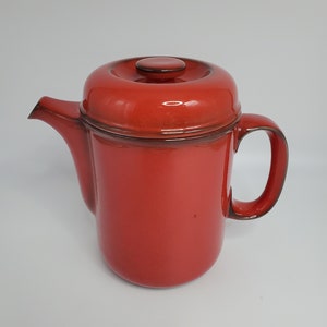 May include: A red ceramic teapot with a lid. The teapot has a rounded body and a handle on the right side.