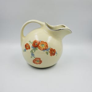 May include: A cream-colored ceramic pitcher with a rounded body and a curved handle. The exterior features a floral pattern of orange poppies and small blue flowers with green stems. The rim of the pitcher is accented with a thin silver metallic trim.