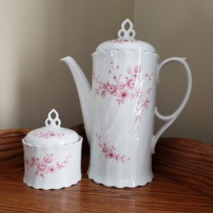 May include: White porcelain coffee pot and sugar bowl with a pink floral pattern. The coffee pot has a fluted design and a lid with a knob. The sugar bowl has a lid with a knob.
