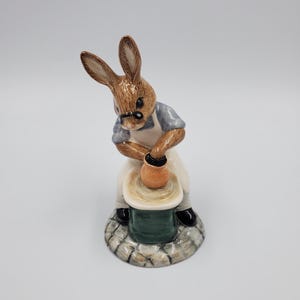 Peut inclure: Figurine en céramique d'un lapin brun portant des lunettes, une chemise bleue et un tablier blanc, travaillant à un tour de potier. Le lapin est concentré sur la mise en forme d'un petit pot orange. La base de la figurine a une texture de pierre.
