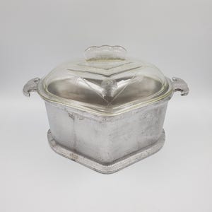 May include: A silver aluminium casserole dish with a glass lid. The lid has a decorative pattern of triangles and circles. The dish has two handles on the sides.