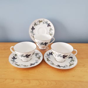 May include: Three white teacups with matching saucers. Each piece features a delicate leaf pattern in shades of blue, green, and grey, with a silver trim. The teacups and saucers are arranged on a wooden surface.