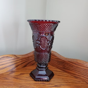May include: A deep red glass vase featuring a flared rim and a textured diamond pattern across the body. The vase has a central band with embossed oval shapes and sits on a solid, hexagonal base with a scalloped edge at the top and bottom.