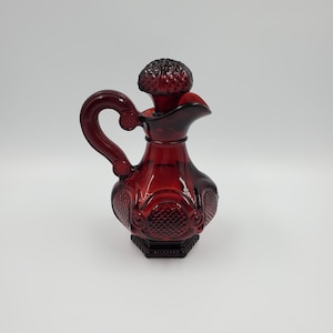 May include: A deep red glass decanter featuring a textured diamond pattern on the body and a matching stopper. The vessel has a curved handle with circular detailing and a wide pouring spout, showcasing a translucent finish and a hexagonal base.