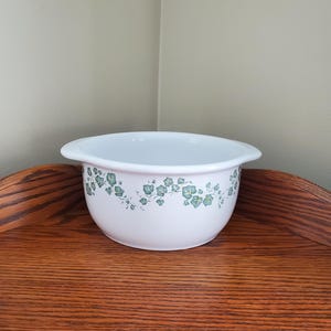 May include: White ceramic bowl with a green floral pattern around the rim. The bowl has a rounded shape with a slight lip and is sitting on a wooden surface. The floral design features small green leaves and flowers.