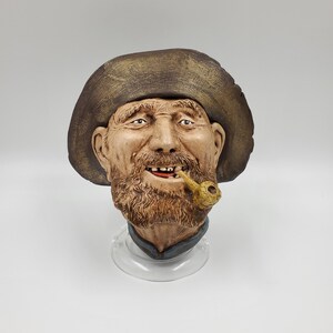 May include: A ceramic sculpture of a man's head wearing a brown hat, with a beard and a pipe in his mouth. The sculpture is painted in realistic colours and has a detailed expression.