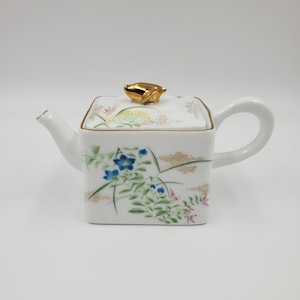 May include: White porcelain teapot with a square body and a curved handle. The lid is topped with a gold-coloured frog-shaped knob. Floral designs in blue, green, and gold decorate the teapot's surface.