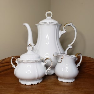 May include: A white porcelain tea set with gold trim. The set includes a teapot, creamer, and sugar bowl. The teapot has a classic design with a rounded body and a curved spout. The creamer and sugar bowl are both small and have a simple design.
