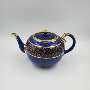 Peut inclure: Théière en céramique bleue et dorée. Le corps est rond et possède un couvercle. Elle présente un motif moucheté bleu et marron, avec des bordures dorées sur le couvercle, la poignée et le bec. La poignée est incurvée.