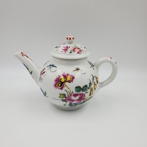 May include: A white porcelain teapot with a floral design. The teapot has a round body and a spout on the left side. The lid is decorated with a floral design and has a small knob on top.