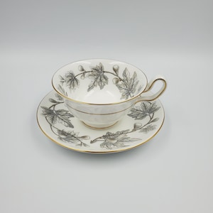 May include: A white teacup and saucer set with a gold rim and a black and gray leaf design. The teacup has a curved handle and sits on a matching saucer. The set is designed for tea service.