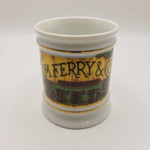May include: White ceramic mug with a gold rim and a yellow, green, and brown graphic of the D.M. Ferry & Co. Celebrated Seeds logo.