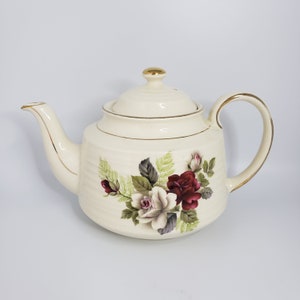 May include: A white teapot with a gold trim and a floral design featuring red and white roses. The teapot has a lid with a gold knob.