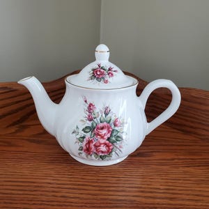 May include: White porcelain teapot with a floral design featuring pink roses and green leaves. The teapot has a gold trim around the lid and spout.