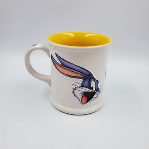 May include: White ceramic mug featuring Bugs Bunny with a yellow interior. The mug has a handle and a cartoon image of Bugs Bunny's face. The mug is a standard size for coffee or tea.