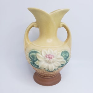 May include: A yellow ceramic vase with two handles and a white flower with pink accents. The vase has a brown base and a green leaf design.