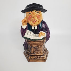 May include: A ceramic figurine of a person wearing a black hat and purple robe, holding a book and standing on a wooden base. The figurine is painted with realistic details, including facial features and clothing.