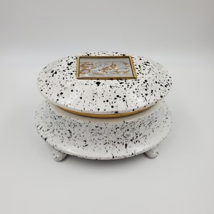 May include: A white ceramic trinket box with black splatter paint and a gold trim. The lid features a silver and gold embossed design inside a gold frame.