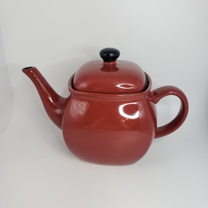 May include: A red ceramic teapot with a black knob on the lid. The teapot has a rounded body and a square lid.