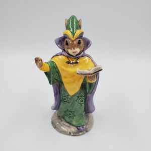 Peut inclure: Figurine en céramique représentant une souris habillée en magicien, vêtue d'une robe verte et jaune avec une cape et un chapeau violets. La souris tient un livre et se tient sur une base grise. Thème fantastique.