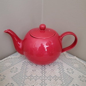 Peut inclure: Une théière rouge vif avec un couvercle et une anse assortis. La théière est ronde avec un bec verseur incurvé et repose sur une nappe en dentelle blanche. Le couvercle a un petit bouton rond.