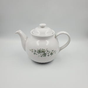 May include: A white ceramic teapot with a lid and handle. The teapot has a green and grey floral pattern around the body. The spout is on the left, and the handle is on the right.