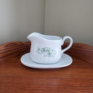 May include: White ceramic creamer and saucer set. The creamer has a handle and a spout, and is decorated with a green floral pattern. The saucer is round with a ridged edge. The set is displayed on a wooden surface.