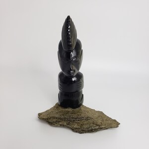 May include: A black carved sculpture of a stylized human figure with a mohawk-like hairstyle. The sculpture is placed on a rough, gray stone base against a white background. The figure has a smooth, polished finish.