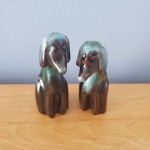 May include: Two ceramic dog figurines, one black and green and the other brown and green, sitting on a wooden surface.