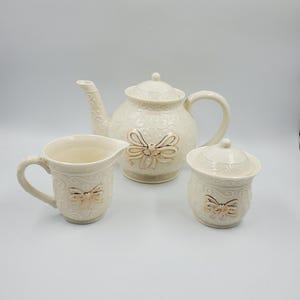 May include: A cream-coloured ceramic tea set consisting of a teapot with a lid, a creamer jug, and a lidded sugar bowl. Each piece features a raised, embossed swirl pattern and a prominent gold-trimmed bow detail on the front surface.