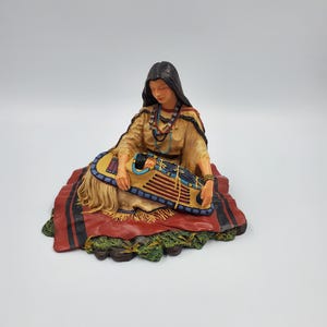 May include: A figurine of a person with long dark hair wearing a tan fringed garment, sitting on a red and black blanket. The person holds a small cradleboard containing an infant, decorated with blue, red, and yellow geometric patterns.