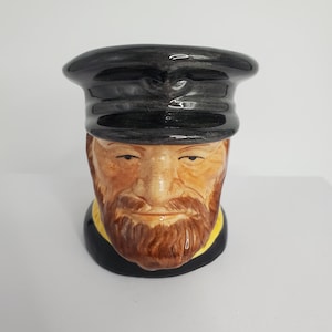 May include: A ceramic mug shaped like a man's head wearing a black cap. The man has a brown beard and moustache. The mug is painted with realistic details.