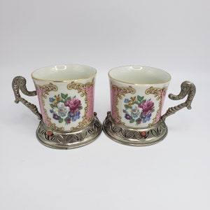 May include: Two porcelain teacups with floral designs and silver accents. The cups have a pink background with a floral pattern in shades of pink, purple, and white. The cups are sitting on a white surface.