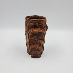 May include: A brown, textured golf bag-shaped container. The container has a detailed design with visible stitching and a small pocket on the front. The top rim is also detailed with stitching and a buckle closure. The container is made of a material that resembles leather.